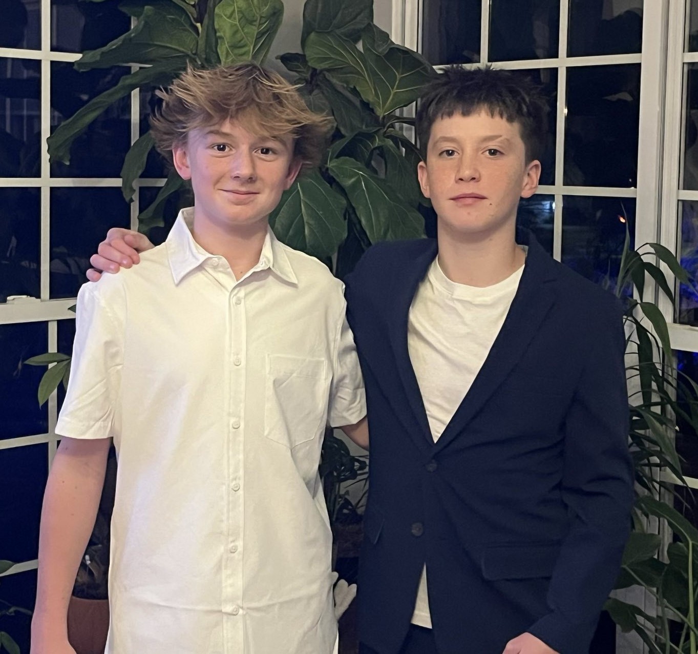 Two teens standing together indoors in front of windows and plants, dressed nicely and smiling